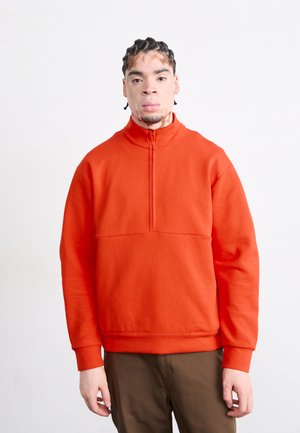 Orange pullover sweatshirt med høj krave, kvart-lynlås foran og en struktureret overflade. Har en lomme foran og ribbede manchetter.