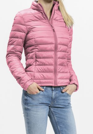 Personne portant une veste matelassée rose zippée, les mains dans les poches et un jean bleu clair sur un fond blanc uni.