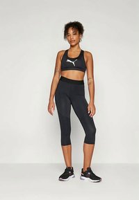 Musta urheiluasu, johon kuuluu valkoisella logolla varustettu lyhyt toppi ja siihen sopivat capri-legginssit. Silkkinen, joustava kangas ja vartalonmyötäinen muotoilu.