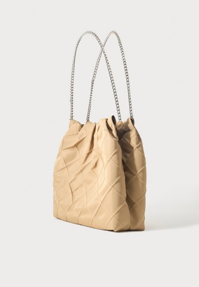 Μπεζ καπιτονέ τσάντα με απαλή, αφράτη υφή και δύο ασημένιες αλυσίδες ως ιμάντες ώμου, εμφανισμένη σε όρθια θέση σε απλό φόντο.