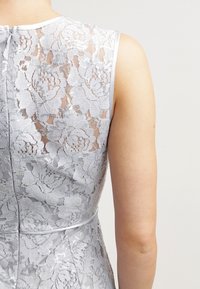 Ärmlös ljusgrå klänning med blommig spets och paljett detaljer, visad från baksidan med en dragkedja och en figurnära midja.