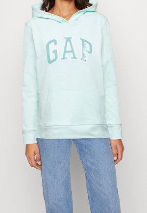 Femme portant un sweat à capuche GAP bleu clair avec poche avant et un jean bleu, debout devant un fond uni clair.
