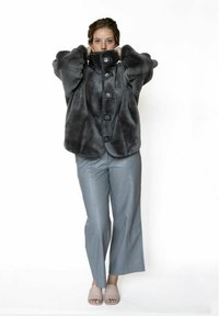 Grijze jas van nep bont met oversized mouwen, hoge kraag en zwarte knopen, gecombineerd met grijze leren broek en lichtgekleurde sandalen.