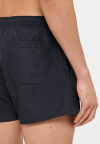 Shorts de bain noirs en tissu léger, dotés d'une taille élastique et d'une poche arrière unique. Texture lisse, coupe au-dessus du genou.