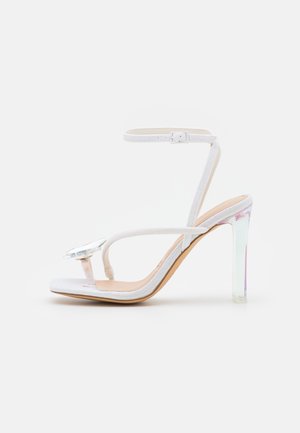Sandales à talons hauts pailletées blanches avec un talon bloc transparent, dotées d'une seule bride de cheville et d'un grand bijou décoratif sur le devant.