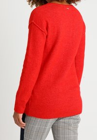 Pull en tricot rouge avec un col rond et des poignets côtelés. Texture douce avec une coupe légèrement ample, associé à un pantalon à carreaux.