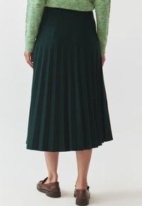 Gonna a pieghe al ginocchio di un verde scuro, realizzata in tessuto textured. Presenta pieghe verticali e una cintura liscia. Scarpe marroni completano il look.