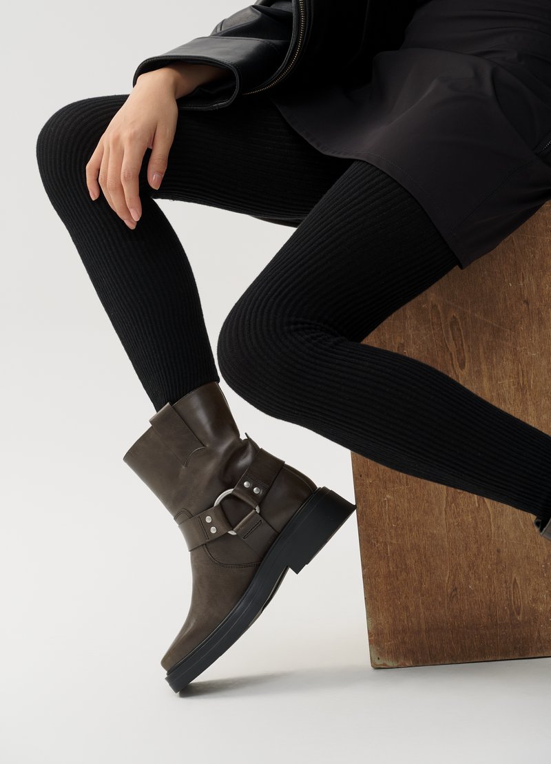 Braune Lederstiefelette mit einem breiten Riemen und einer Metall-Schnalle. Verfügt über eine klobige schwarze Sohle und markante Nähte. Getragen mit schwarzen gerippten Strumpfhosen.