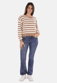 Maglione fluffy a righe marroni e bianche con scollatura rotonda. Abbinato a jeans a zampa d'elefante blu e sneakers bianche, in piedi di fronte a uno sfondo semplice.