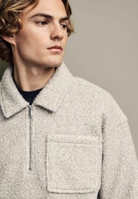 Giacca in pile grigio chiaro con mezza zip, tasca visibile, tessuto texturizzato e collo abbottonato. Design semplice e pratico con una finitura morbida.