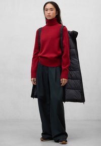 Červený pletený svetr s vysokým límcem, v kombinaci s volnými tmavě zelenými kalhotami a černou vatovanou bundou přehozenou přes rameno.