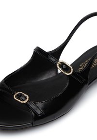 Scarpa in vernice nera con punta chiusa, lati aperti e cinturino alla caviglia. Presenta dettagli in metallo dorato con strass.