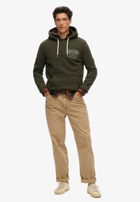 Groene hoodie met trekkoorden en een voorzak, gelaagd over een geruiten shirt. Gecombineerd met beige broek en witte sneakers.