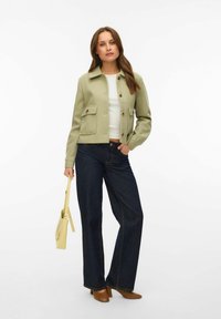 Femme portant une veste vert clair, un haut blanc, un jean large foncé, des chaussures marron, tenant un petit sac à main jaune sur un fond blanc.