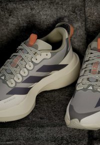 Ζευγάρι γκρι και λευκά αθλητικά παπούτσια Adidas με πορτοκαλί λουράκια και χοντρές σόλες, τοποθετημένα σε μια σκοτεινή υφή.
