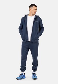 Dunkelblaues Hoodie und Jogginghose aus weichem Material, mit einem Reißverschluss vorne, gerippten Bündchen und passenden Turnschuhen für einen abgestimmten Look.