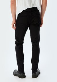 Vaqueros de denim negros con corte ajustado, que cuentan con dos bolsillos traseros y una textura suave. Llevados con zapatos de tacón negros sobre un fondo claro.