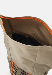 Beige batoh s otevřeným vrchem a oranžovým zipem. Obsahuje černou vnitřní podšívku a boční síťovou kapsu, nastavitelné popruhy a dekorativní prvky.