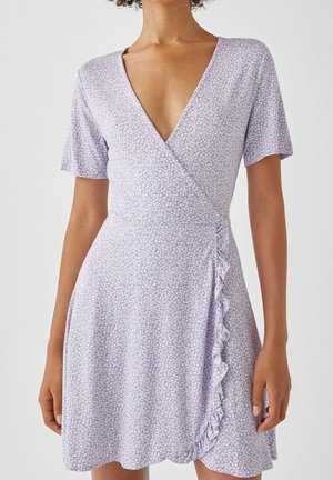 Femme portant une robe portefeuille violet clair avec un petit imprimé floral blanc, manches courtes, décolleté en V profond et un détail volanté sur le côté.
