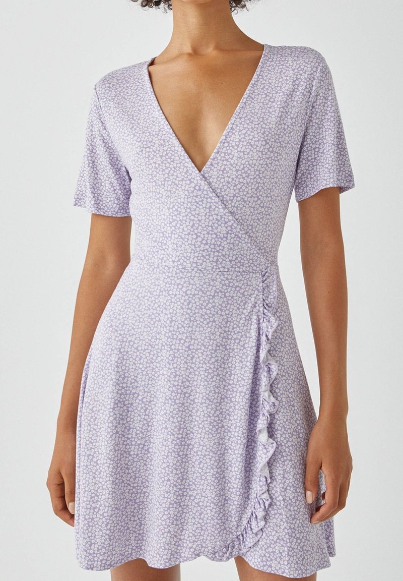 Femme portant une robe portefeuille violet clair avec un petit imprimé floral blanc, manches courtes, décolleté en V profond et un détail volanté sur le côté.