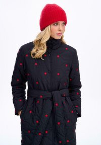 Čierny prešívaný kabát s červenými bodkami, vysokým golierom a opaskom, s dlhými rukávmi. Skombinovaný s červenou pletenou čiapkou.