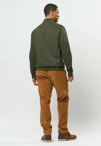 Homme debout de dos, portant une veste vert foncé, un pantalon marron et des bottes en cuir marron sur un fond clair uni.