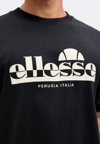 Μαύρο μπλουζάκι με λευκό λογότυπο "ellesse" και κείμενο "Perugia Italia" στο στήθος, φορεμένο από ένα άτομο με ορατό λαιμό και κάτω μέρος προσώπου.