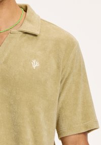 Polo tričko vo svetlej olivovej terakotovanej látke, s klasickým golierom a malým bielym vyšitým logom na ľavej strane.