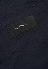 Marineblauer, strukturierter Stoff mit matter Oberfläche, versehen mit einem schwarzen Etikett, auf dem weißer "Matinique" Schriftzug aufgenäht ist. Schlichte, rechteckige Form mit minimalen Akzenten.