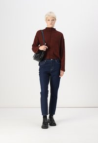 Bordo aukštu kaklu megztinis, tamsiai mėlyni siauri džinsai ir juodi auliniai batai. Modelis laiko mažą juodą rankinę, minimalizmo dizainas.