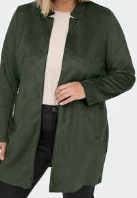 Abrigo largo de ante verde con cuello y bolsillos laterales, con un corte recto y diseño minimalista, combinado con una blusa clara y pantalones negros.