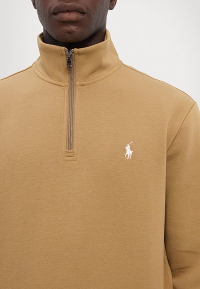 Khaki polar z zamkiem w kolorze białym, wykonany z gładkiego materiału; posiada niewielkie białe logo na piersi oraz zamek w kołnierzu do regulacji dopasowania.