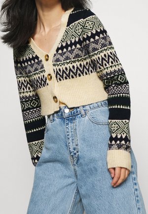 Femme portant un cardigan court en maille à motif avec des boutons en bois et un jean denim bleu clair taille haute, sur un fond uni.