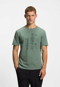 T-shirt di cotone in verde tenue con maniche corte, colletto tondo e stampa grafica nera che presenta forme astratte di edifici.