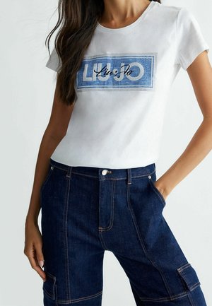 Mujer con camiseta blanca con logo azul "Liu Jo" y pantalones vaqueros de talle alto azul oscuro con bolsillos laterales, con la mano apoyada en el bolsillo.