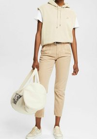 Sweatshirt à capuche beige sur un t-shirt blanc, associé à un pantalon tapered marron clair et des baskets beiges. Un sac rond beige avec un logo.