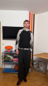 Jeune homme en sweat à capuche noir et blanc et pantalon noir se tient dans une pièce avec une télévision, une lampe orange, un ballon de basket et une valise argentée décorée d'autocollants.