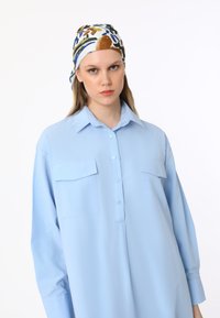 Jasnoniebieska koszula z długim rękawem, posiadająca dwie kieszenie na piersi i zapinana na guziki. Model nosi wzorzysty szalik w kolorach białym, niebieskim i brązowym.