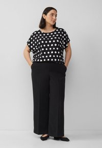 Vrouw in een zwarte blouse met witte stippen en zwarte wijde broek, staande met haar handen in de zakken, kijkend naar opzij.