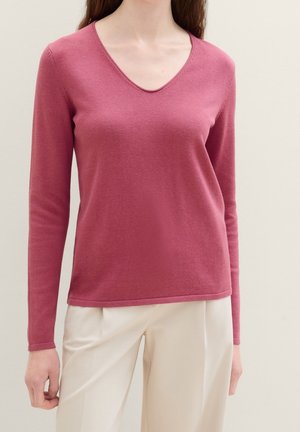 Femme portant un pull à manches longues couleur rose poussiéreux avec un col en V et un pantalon ample blanc cassé, posant devant un fond uni.