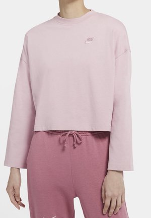 Osoba nosząca jasnoróżową krótką bluzę Nike z długim rękawem oraz ciemnoróżowe spodnie dresowe ze ściągaczem, stojąca na jednolitym tle.