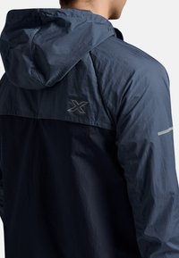Marine lichtgewicht jack met capuchon, gemaakt van een gladde, gestructureerde stof. Reflecterende accenten langs de schouder en logodetail op de achterkant.