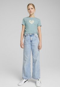 Camiseta ajustada de color azul claro con un diseño de corazón en llamas color crema, combinada con jeans de tiro alto y pierna ancha de lavado claro y zapatillas blancas.