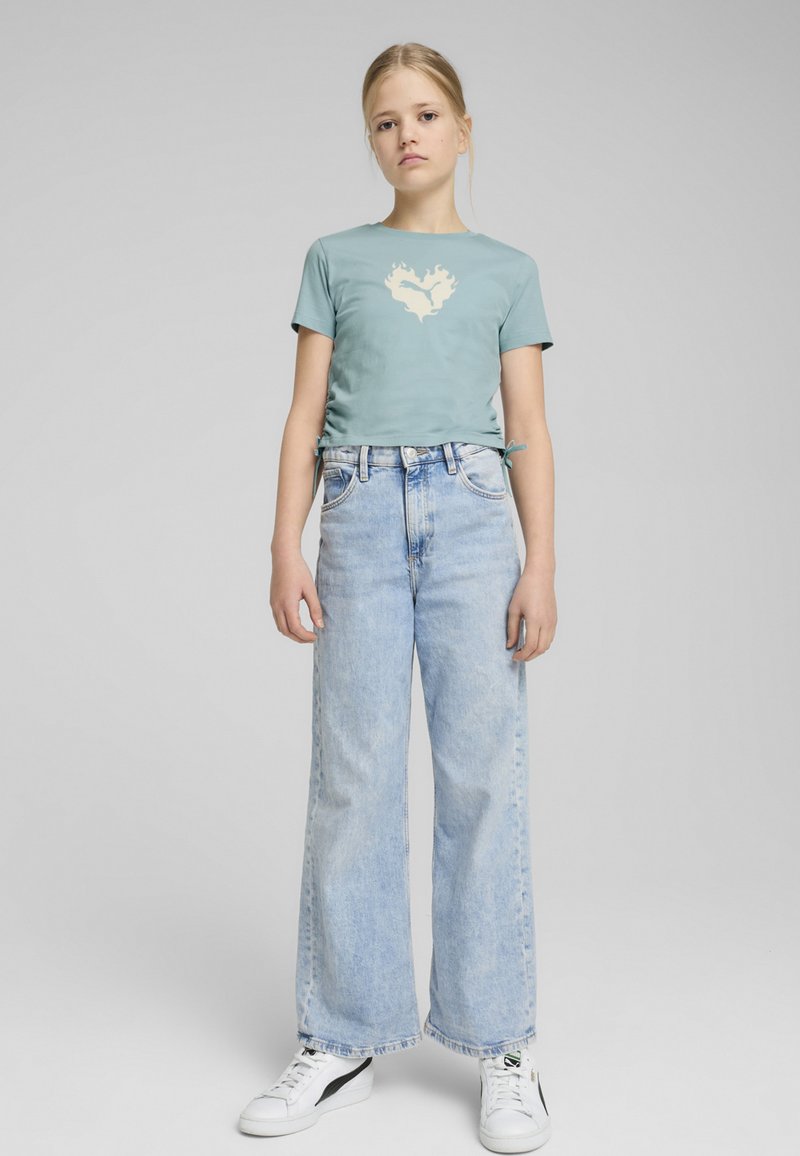 Camiseta ajustada de color azul claro con un diseño de corazón en llamas color crema, combinada con jeans de tiro alto y pierna ancha de lavado claro y zapatillas blancas.