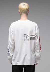 Άσπρο πουκάμισο με μακριά μανίκια, με έντονα τυπωμένα κείμενα σε κόκκινο και μπλε στα μανίκια, γραφιστικό σχέδιο στην πλάτη και άνετη εφαρμογή.