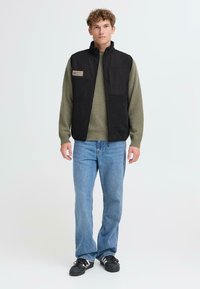Schwarze Fleeceweste mit hohem Kragen, Fronttaschen; getragen über einem olivfarbenen Pullover, kombiniert mit hellblauen Jeans und schwarzen Sneakers.