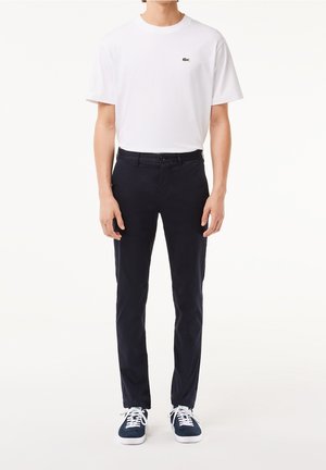 Homme portant un t-shirt blanc à manches courtes avec un petit logo, un pantalon noir coupe slim et des baskets bleu marine avec des lacets blancs, debout devant un fond blanc.