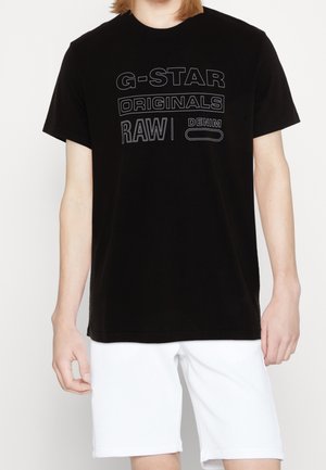 T-shirt noir à manches courtes avec un graphisme frontal indiquant "G-STAR ORIGINALS RAW | DENIM" en blanc et gris. Porté avec un short blanc.
