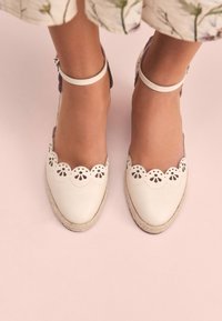 White espadrille shoes with scalloped edges and cutout flower designs. Natural jute sole and leather ankle straps. Flat, rounded toe.