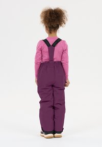 Lila Schneehose mit verstellbaren schwarzen Trägern, gummierten Akzenten und weit geschnittenem Bein. Kombiniert mit einem pinken Langarmshirt.
